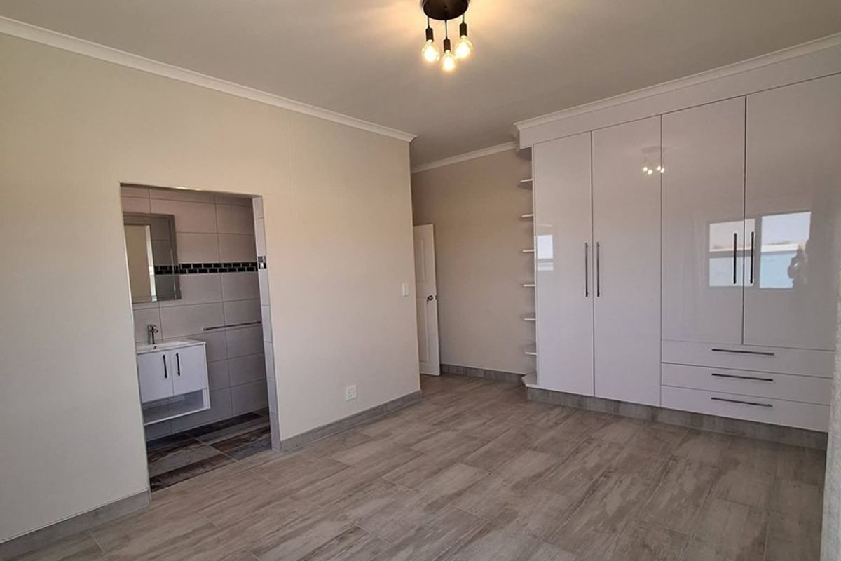 A spacious, modern bedroom featuring neutral tiled flooring, extensive built-in white cabinetry, and an adjoining en-suite bathroom with a vanity.