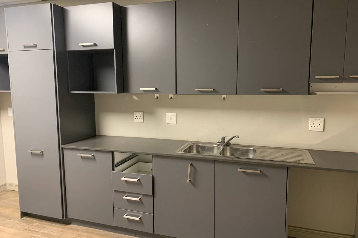 A functional office kitchenette featuring grey built-in cabinetry, a stainless steel sink, and laminate countertops, currently requiring minor repairs to drawer units.