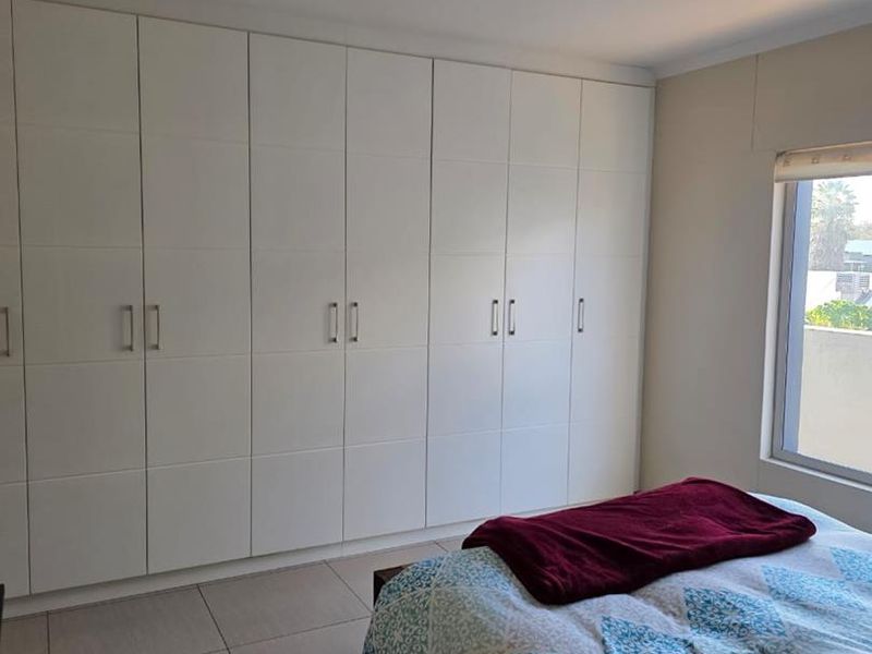 A well-maintained bedroom featuring extensive floor-to-ceiling built-in cupboards, neutral tiled flooring, and natural light from a side window.