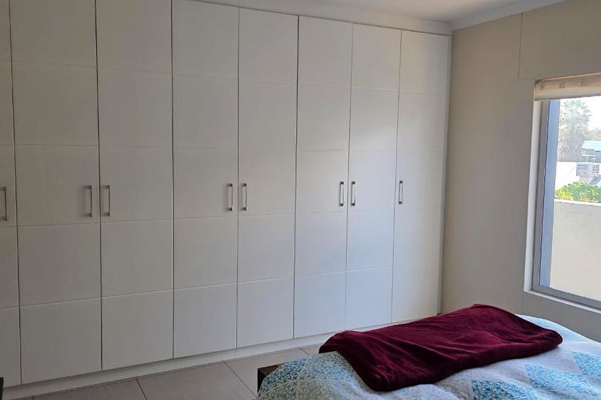 A well-maintained bedroom featuring extensive floor-to-ceiling built-in cupboards, neutral tiled flooring, and natural light from a side window.