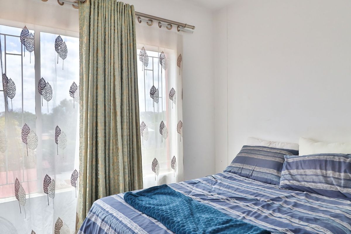 A bright bedroom featuring large windows with security bars and decorative curtains, offering natural light and a view of the surrounding area.