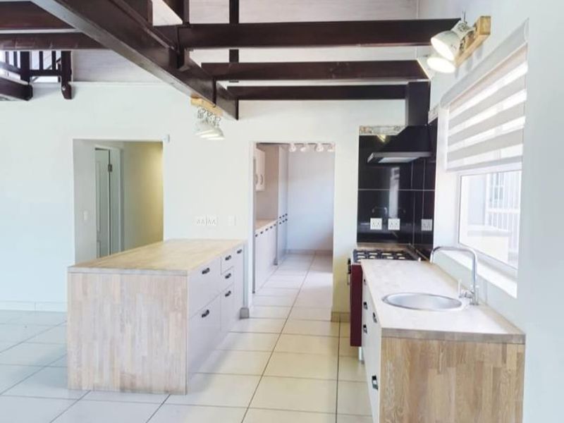 Modern kitchen featuring a central island, timber cabinetry, exposed ceiling beams, and tiled flooring, with an open-plan layout leading to a mezzanine level.