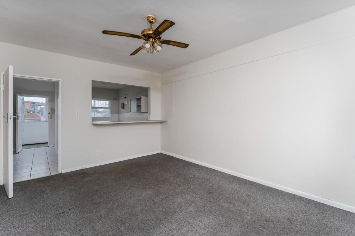 An empty living room featuring a ceiling fan and a convenient serving hatch opening into the kitchen area, with access to a tiled hallway.