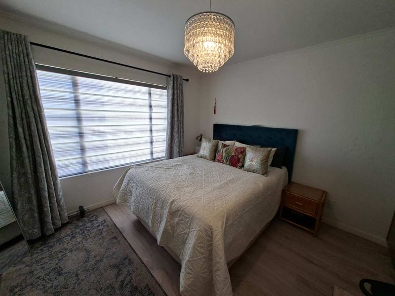 A well-lit bedroom featuring light-toned laminate flooring, a large window with modern blinds, and a decorative ceiling light fixture.