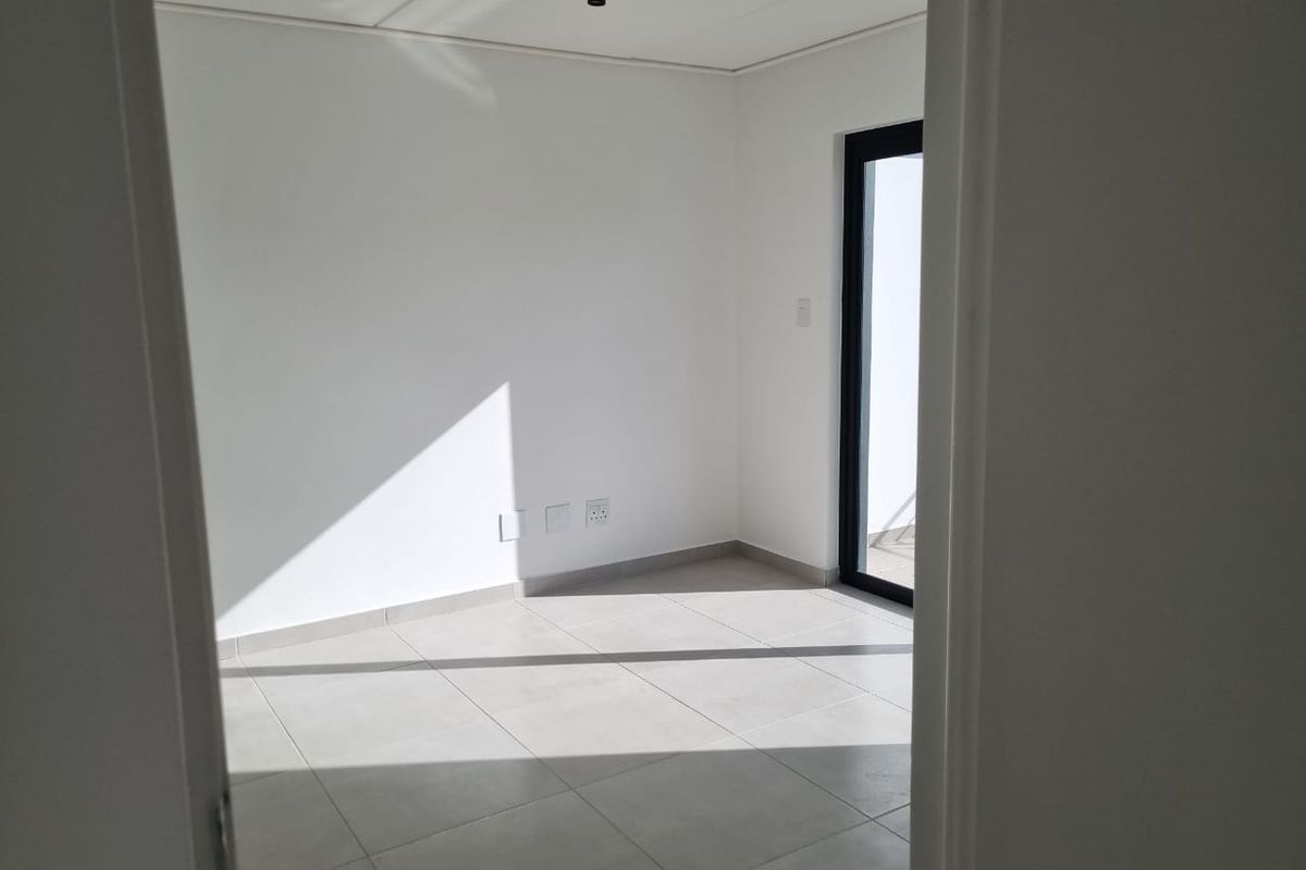 A bright, empty bedroom featuring neutral tiled flooring, modern track lighting, and a glass door providing access to an outdoor area.