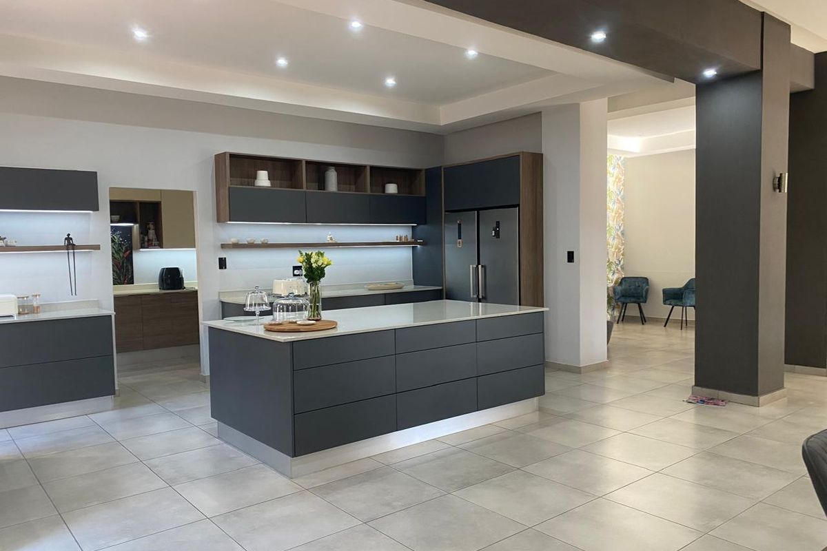 Modern open-plan kitchen featuring a large central island, sleek dark cabinetry, stone countertops, integrated appliances, and recessed lighting, with an adjacent scullery area.