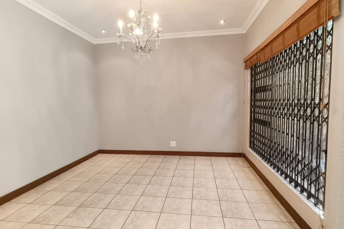 A bright, empty living room featuring neutral tiled flooring, crown molding, and a large window secured with a metal security gate.