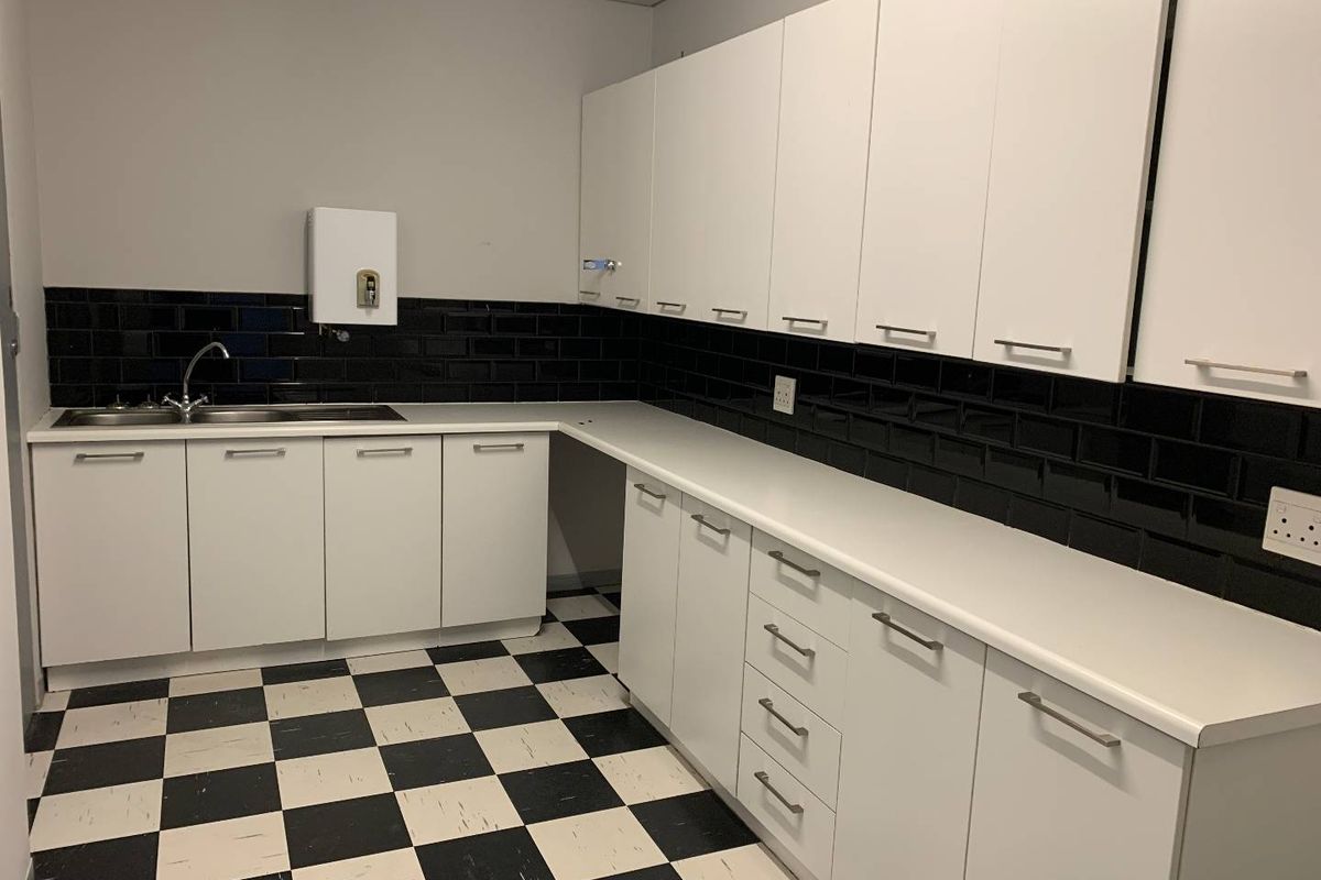 A functional office kitchenette featuring white cabinetry, contrasting black subway tile backsplash, and a durable checkerboard vinyl floor.