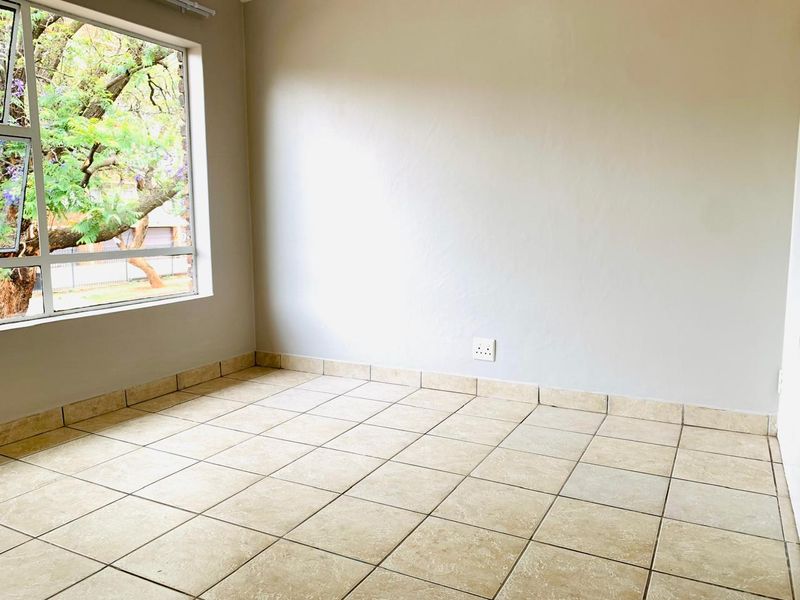 A bright, empty bedroom featuring neutral tiled flooring and a large window providing natural light and a view of the outdoors.