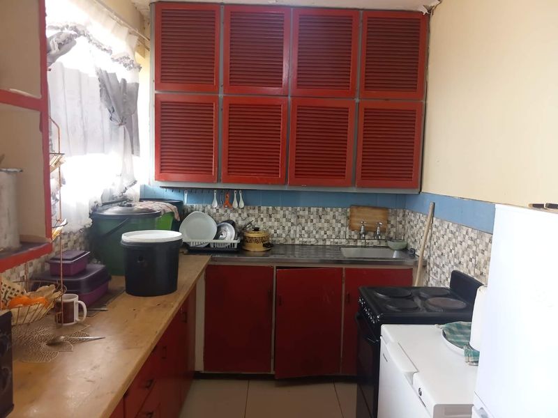 A compact kitchen featuring red louvered cabinetry, a stainless steel sink, and a tiled backsplash, currently showing signs of wear and dated finishes.