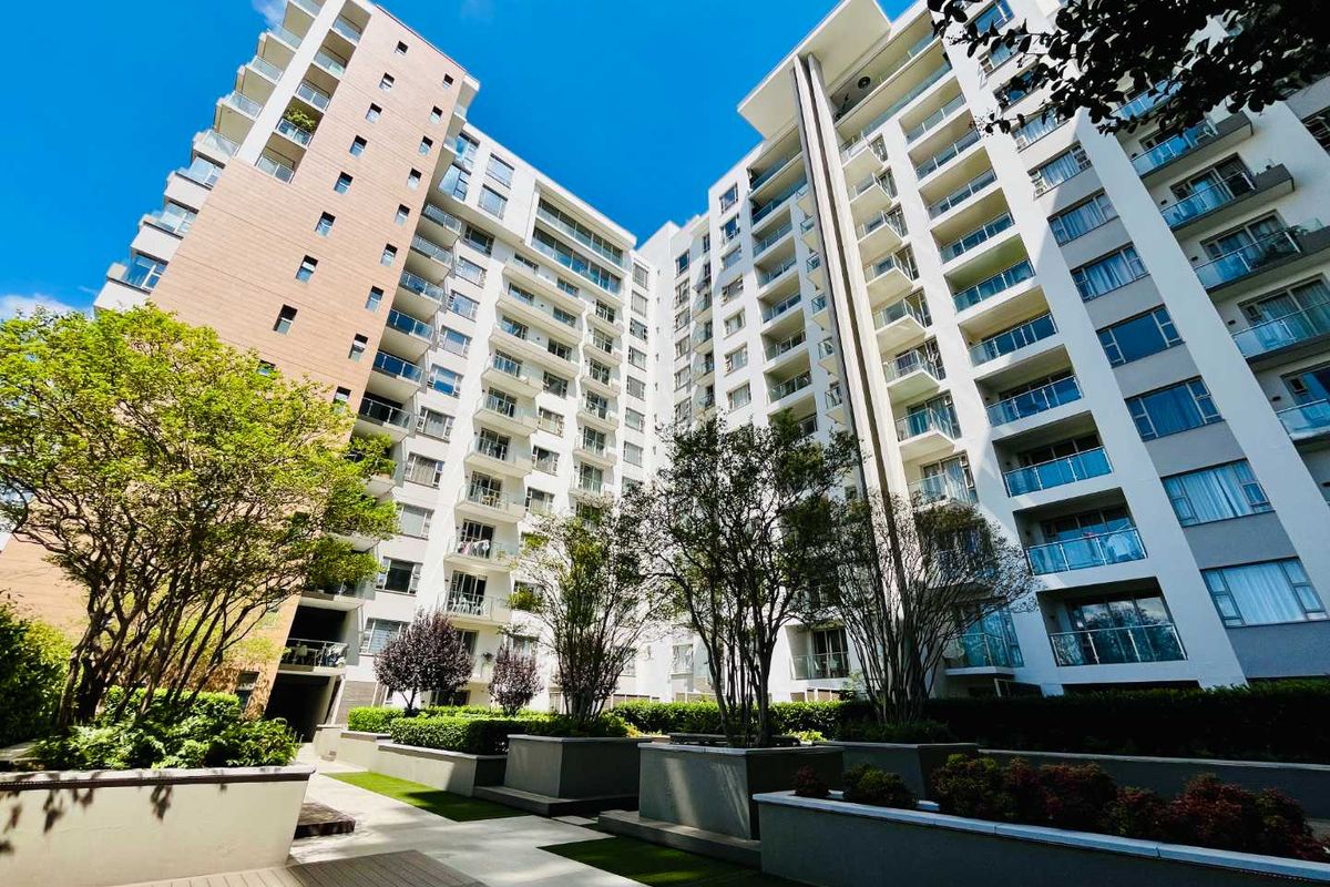 A low-angle view of a modern multi-story apartment complex featuring glass balconies, landscaped garden beds, and paved walkways.