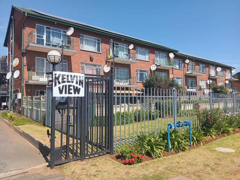 Exterior view of the Kelvin View apartment complex featuring a multi-story brick building with balconies, secure perimeter fencing, and a landscaped front garden area.