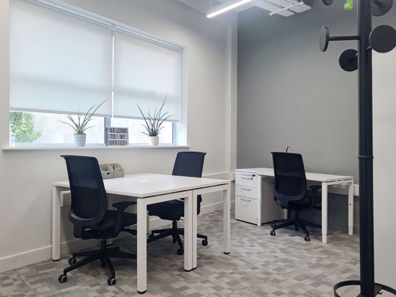 A modern, well-lit office space featuring neutral tones, carpeted flooring, and functional roller blinds, providing a professional and clean working environment.