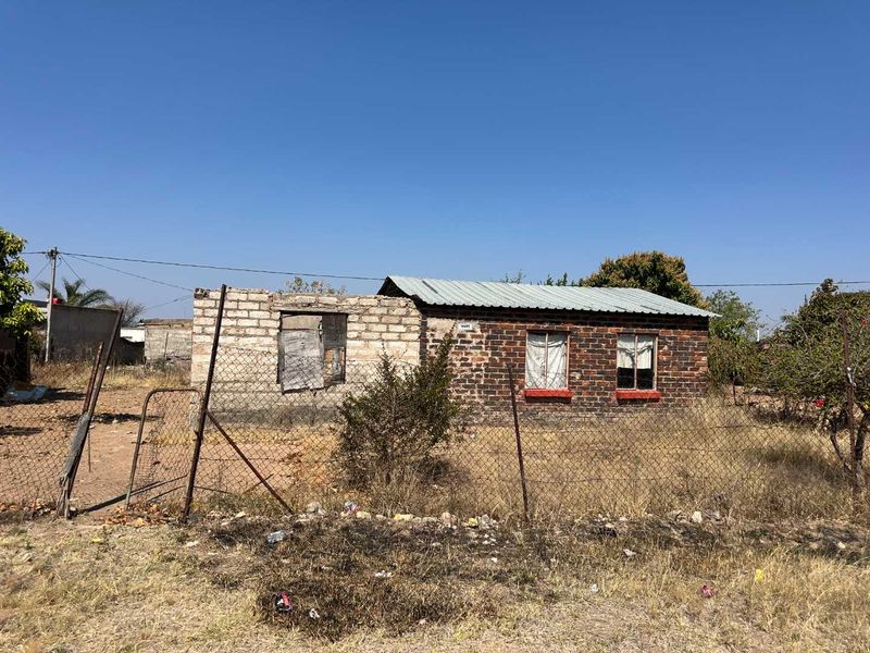 A detached single-story house featuring a brick facade and corrugated metal roof, set on a plot with perimeter chain link fencing.