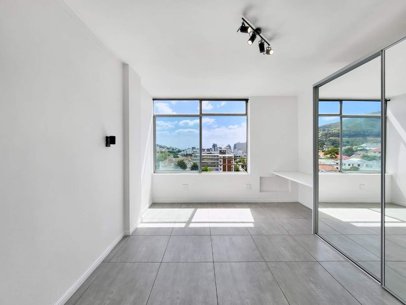 Bright and modern bedroom featuring large windows with city views, tiled flooring, a built-in desk, and a spacious mirrored wardrobe.