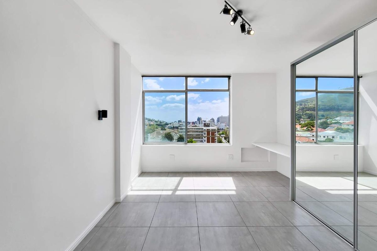 Bright and modern bedroom featuring large windows with city views, tiled flooring, a built-in desk, and a spacious mirrored wardrobe.