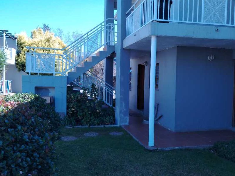 Exterior view of a two-story apartment building featuring an external staircase, balcony with metal railings, and a small garden area.