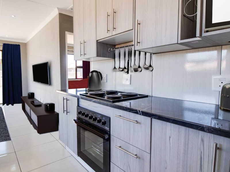 Modern open-plan kitchen featuring light wood-grain cabinetry, dark stone countertops, and integrated appliances, flowing into a bright living area with tiled floors.