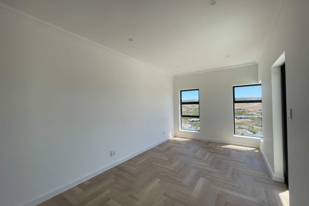 A bright, modern bedroom featuring light-toned herringbone timber flooring, crisp white walls, recessed downlighting, and large windows offering scenic landscape views.