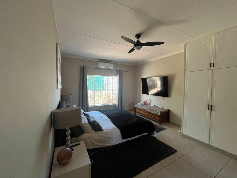 A well-maintained bedroom featuring built-in cupboards, a ceiling fan, and a split-system air conditioner, with tiled flooring and security bars on the window.