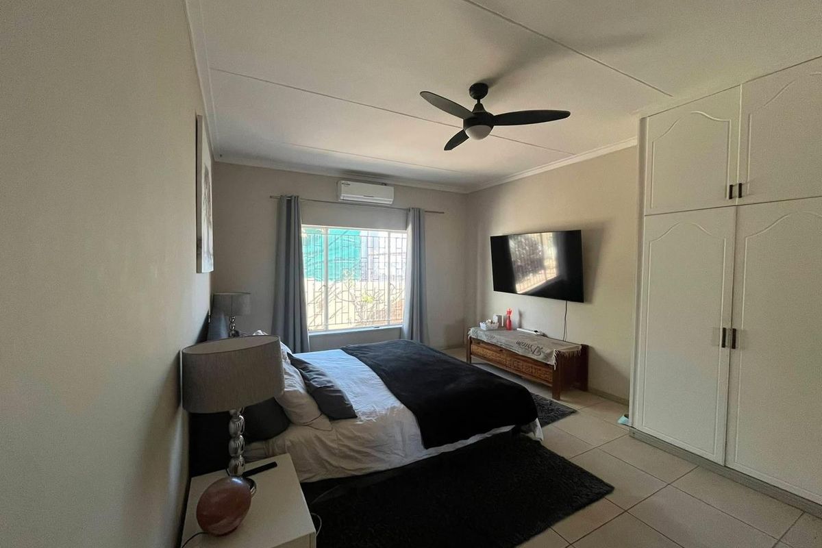 A well-maintained bedroom featuring built-in cupboards, a ceiling fan, and a split-system air conditioner, with tiled flooring and security bars on the window.