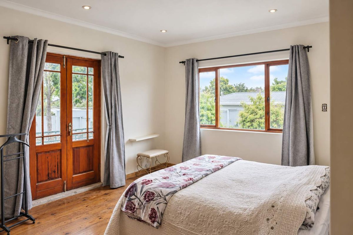 A bright bedroom featuring polished timber flooring, wooden french doors leading outside, and large windows that provide ample natural light.