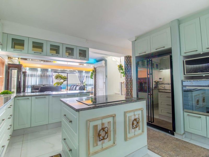 Spacious open-plan kitchen featuring light green cabinetry, granite countertops, a central island with a hob, and integrated appliances, flowing into the living area.