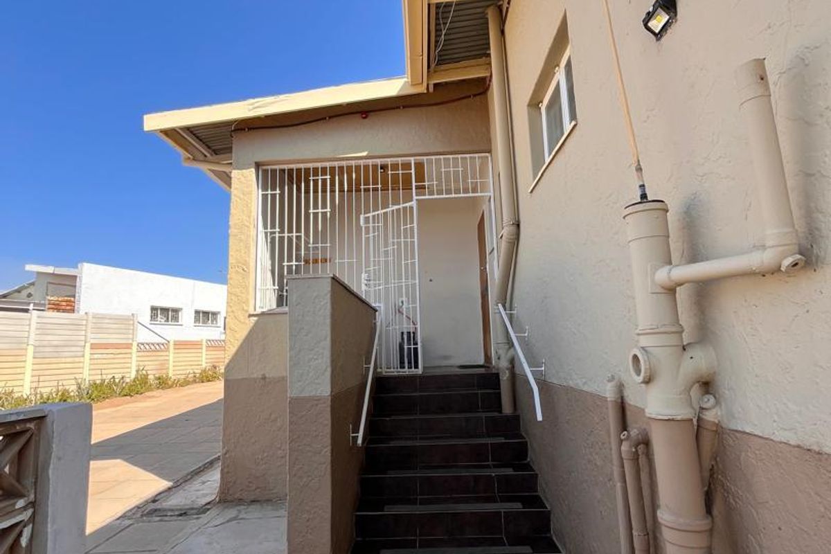 Rear exterior view showing a tiled staircase leading to a secured entrance, featuring external plumbing and paved ground surfaces.