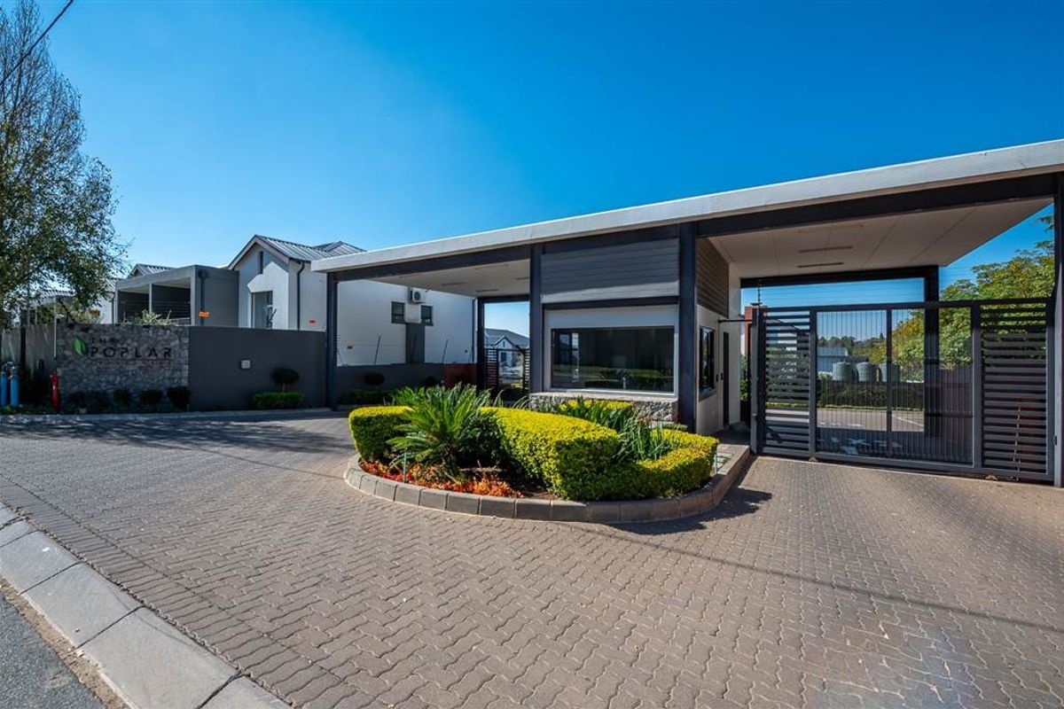 The secure entrance gatehouse of The Poplar apartment complex, featuring modern architectural design, paved driveway, and landscaped garden beds.
