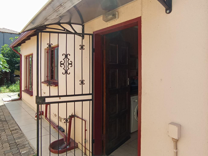 Rear exterior view showing a paved walkway, security gate, and entrance door with visible wear to the exterior wall finish.