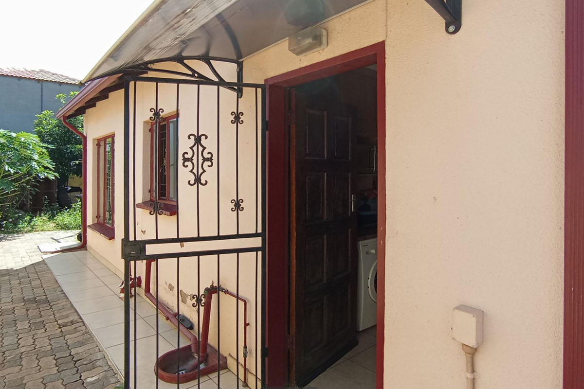 Rear exterior view showing a paved walkway, security gate, and entrance door with visible wear to the exterior wall finish.