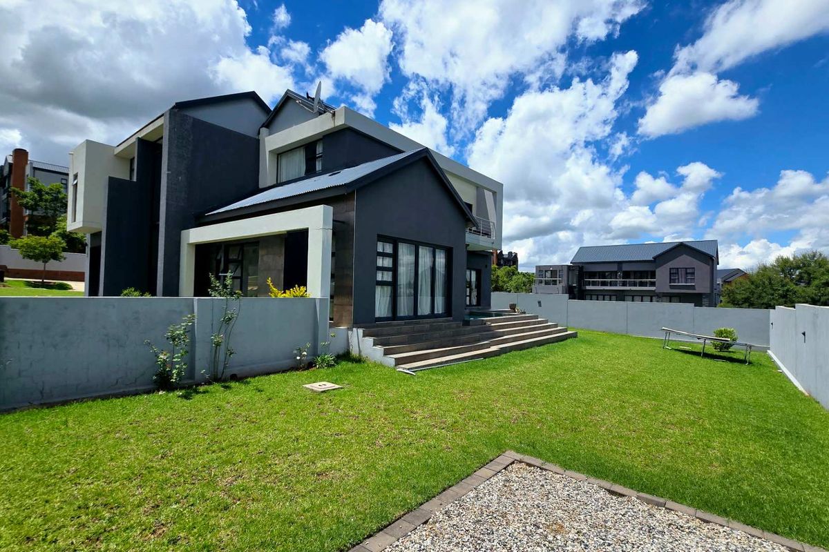 A modern double-story house featuring a dark grey facade, large glass sliding doors, and a spacious rear garden with a paved patio area.