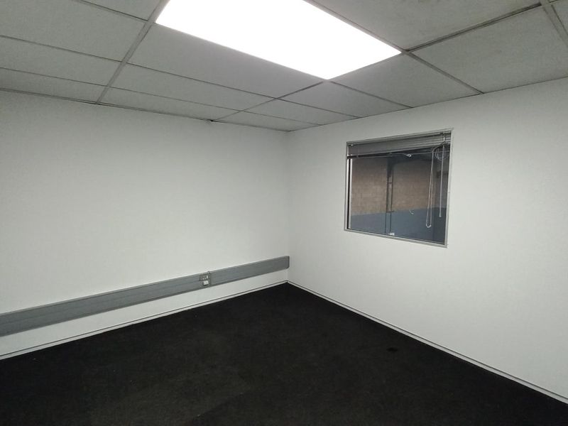 A clean, empty office space featuring a suspended ceiling, recessed lighting, dark carpeting, and a window looking out into the industrial warehouse area.