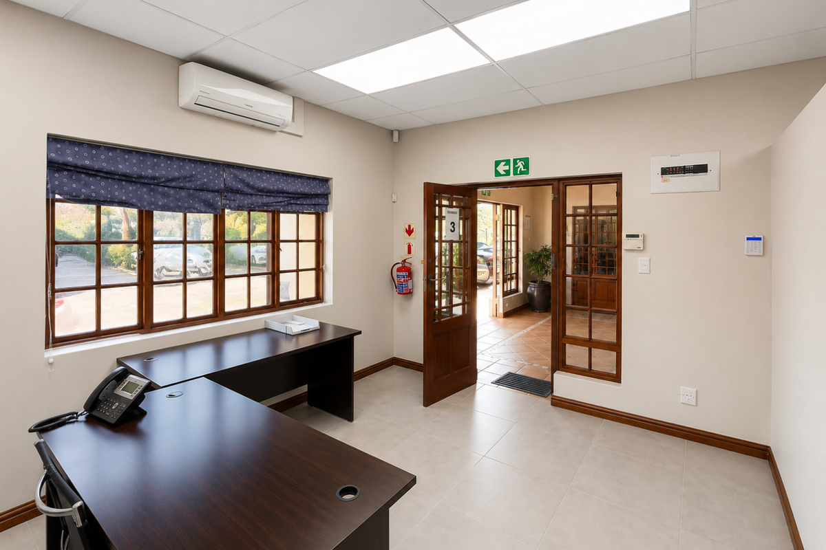 A well-maintained office space featuring tiled flooring, a suspended ceiling, split-system air conditioning, and timber-framed windows providing natural light.