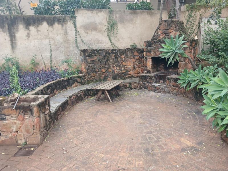 Outdoor entertainment area featuring a rustic stone-built braai, circular brick paving, and integrated stone seating against a garden boundary wall.