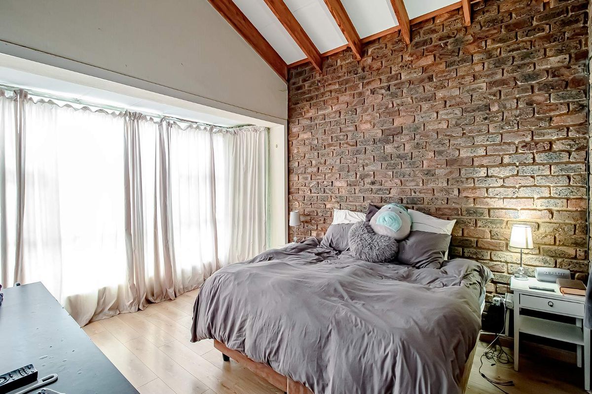 A bedroom featuring a prominent exposed brick feature wall, vaulted ceiling with timber beams, and large windows allowing for ample natural light.