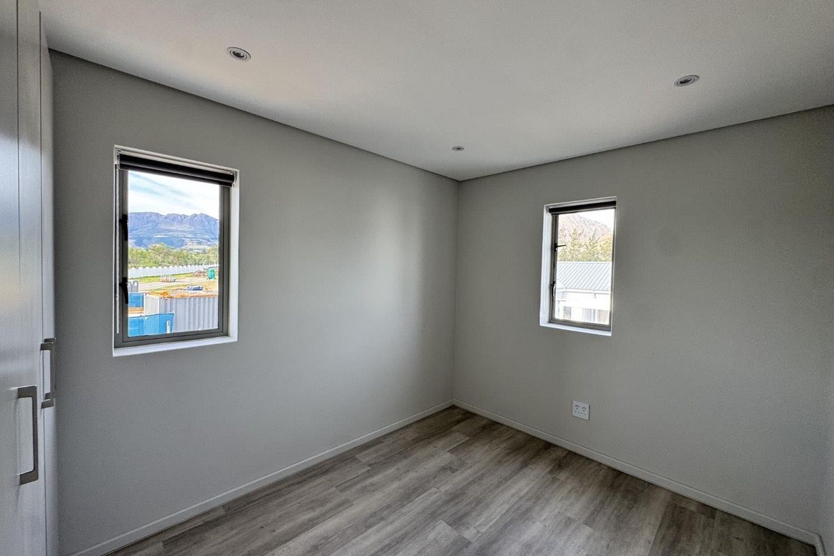 A bright, modern, and empty bedroom featuring neutral walls, laminate flooring, built-in cupboards, and recessed downlights with views through aluminium-framed windows.