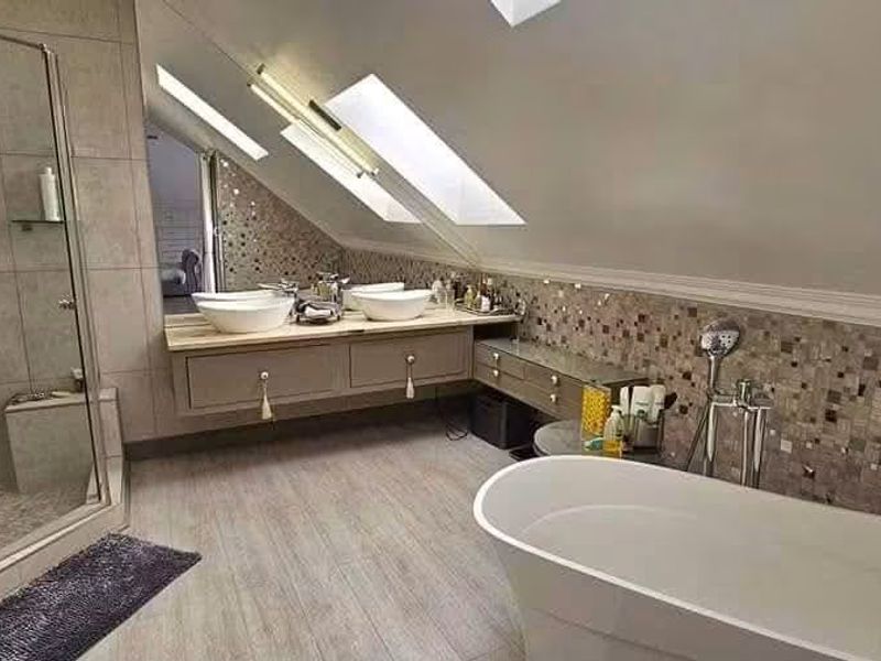 Modern attic bathroom featuring a freestanding bathtub, glass-enclosed shower, dual vessel sinks on a wall-mounted vanity, and natural light from skylights.