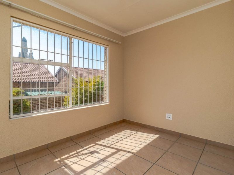 A bright, empty bedroom featuring neutral walls, tiled flooring, and a large window fitted with security bars.