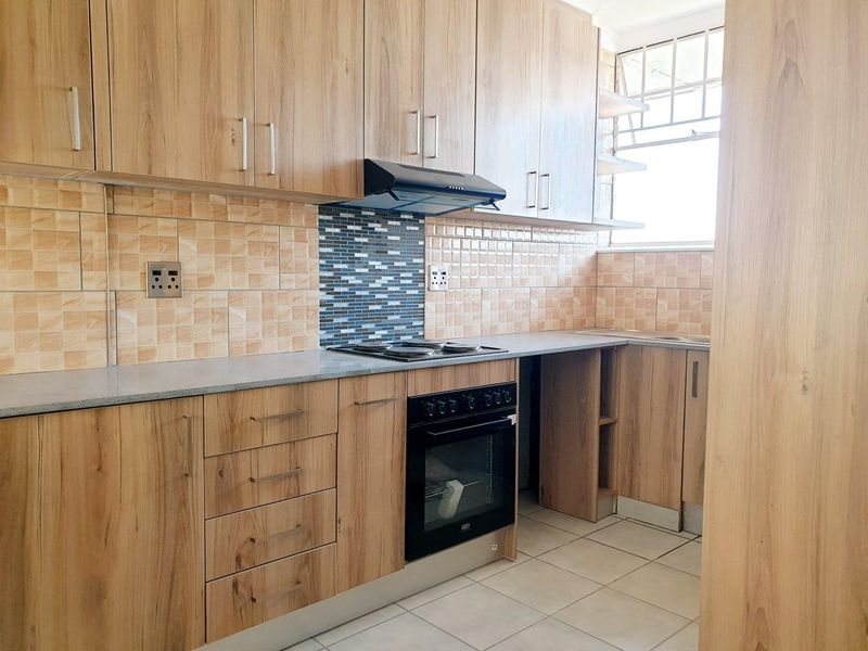 A functional kitchen featuring wood-look cabinetry, laminate countertops, built-in oven, electric hob, extractor fan, and tiled backsplash with security bars on the window.