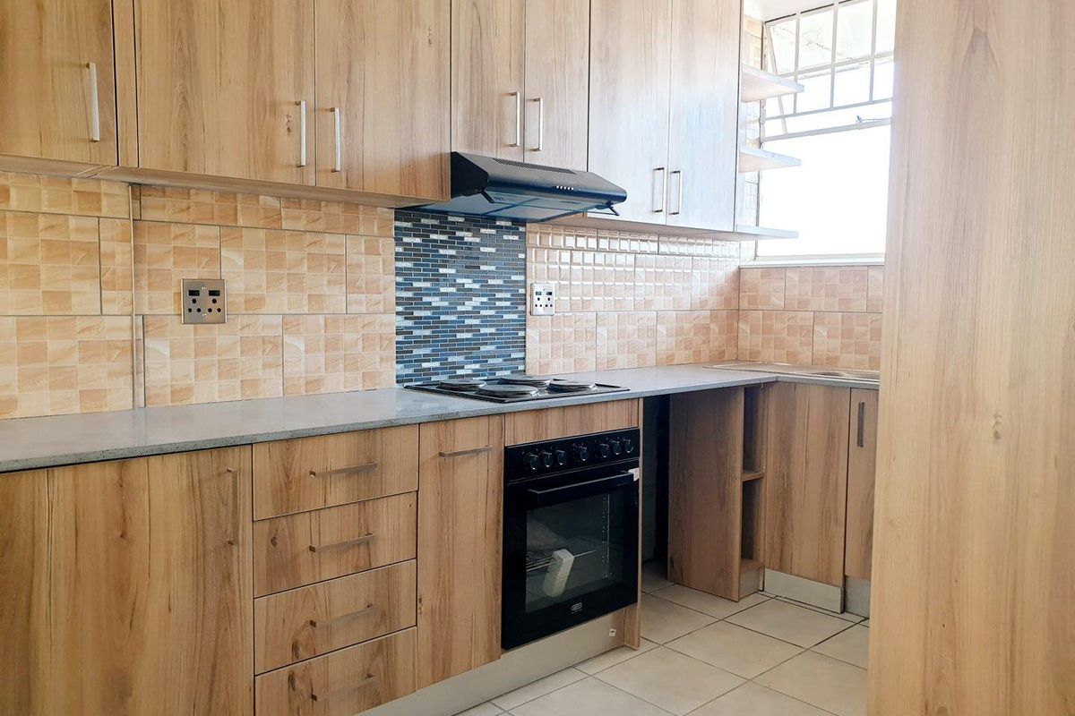 A functional kitchen featuring wood-look cabinetry, laminate countertops, built-in oven, electric hob, extractor fan, and tiled backsplash with security bars on the window.