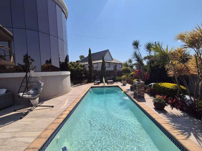 A well-maintained rectangular swimming pool set within a paved patio area, featuring a modern glass-clad building facade and landscaped garden surroundings.