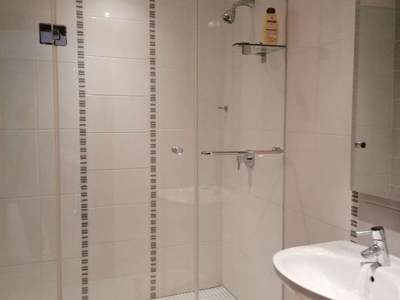A clean, modern bathroom featuring a glass-enclosed shower, pedestal basin with chrome fixtures, and neutral wall and floor tiling.