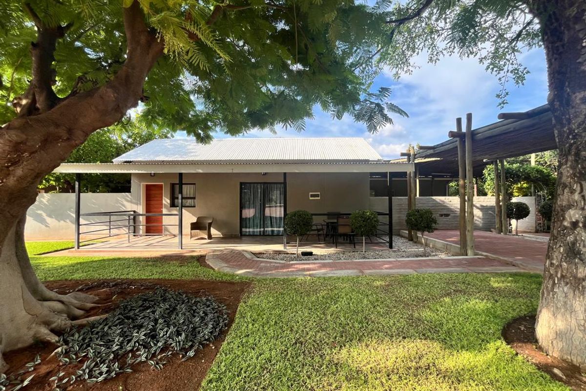 A well-maintained lodge exterior featuring a covered patio, metal roof, and paved walkway set within a landscaped garden with mature trees.