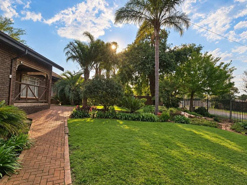 A well-maintained garden featuring a brick paved walkway, lush lawn, mature palm trees, and secure metal perimeter fencing.