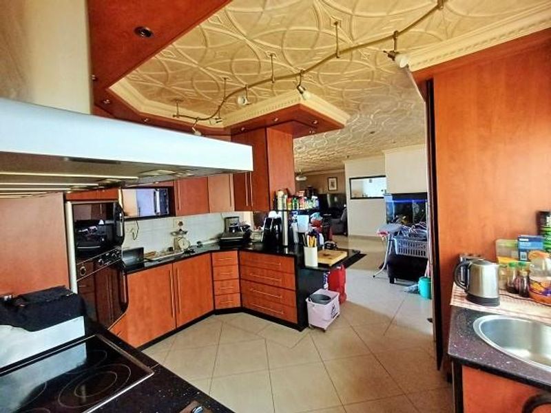 Open plan kitchen featuring wood-finish cabinetry, built-in appliances, and decorative ceiling detail, flowing into an adjacent living area with tiled flooring throughout.