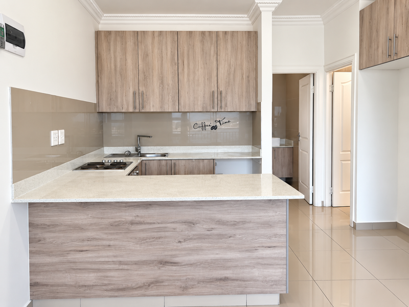 Modern kitchen featuring wood-look cabinetry, stone countertops, integrated hob, and sleek glass splashback, leading into a tiled hallway and adjacent bathroom area.