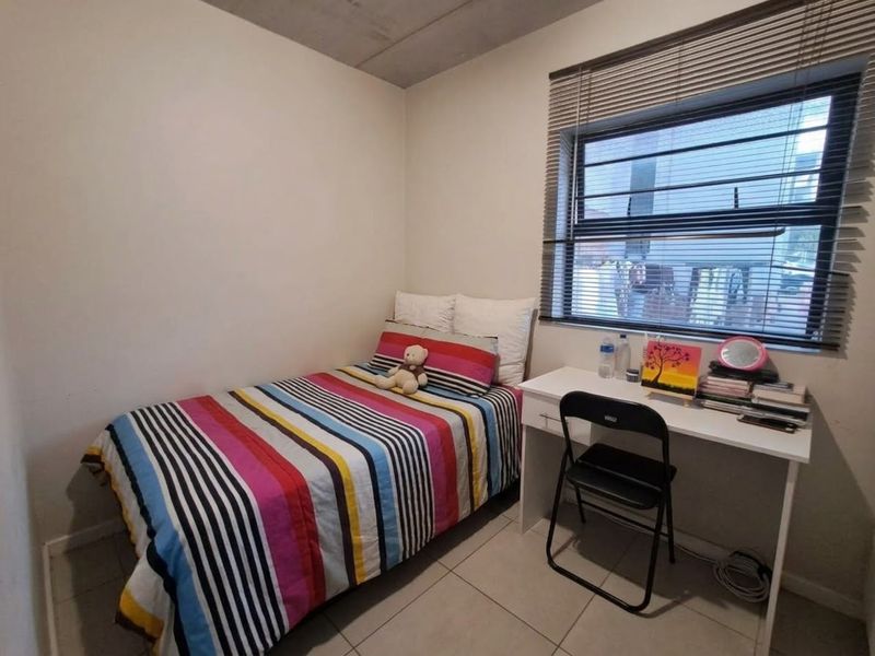 A compact bedroom featuring tiled flooring, an exposed concrete ceiling, and a window fitted with venetian blinds.