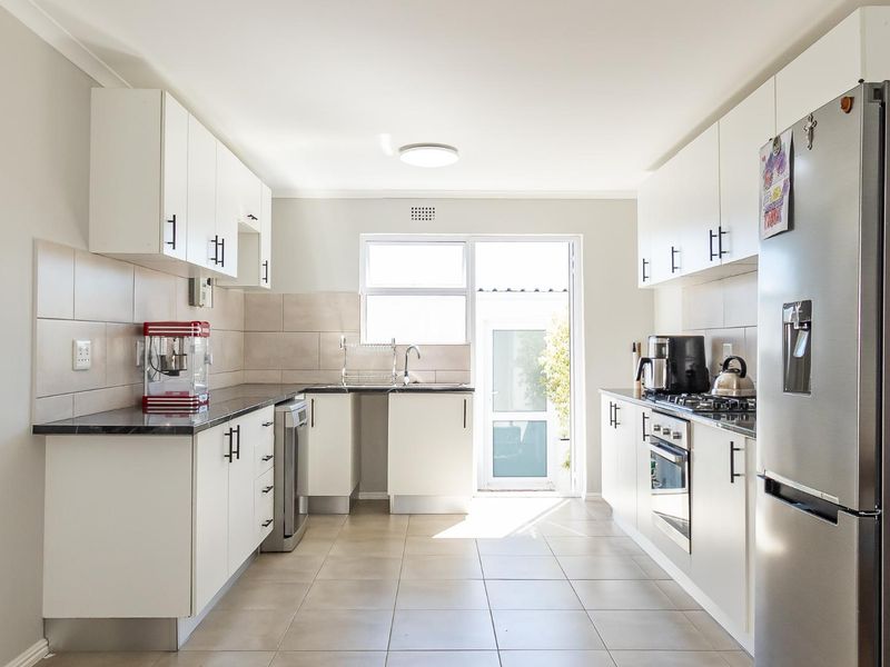 A bright, modern kitchen featuring white cabinetry, dark stone countertops, stainless steel appliances, and tiled flooring with direct access to the rear exterior.