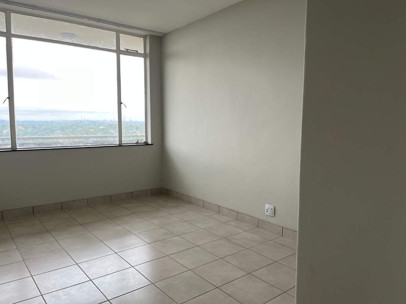 An empty bedroom featuring neutral tiled flooring, large windows with natural light, and a view of the surrounding area.